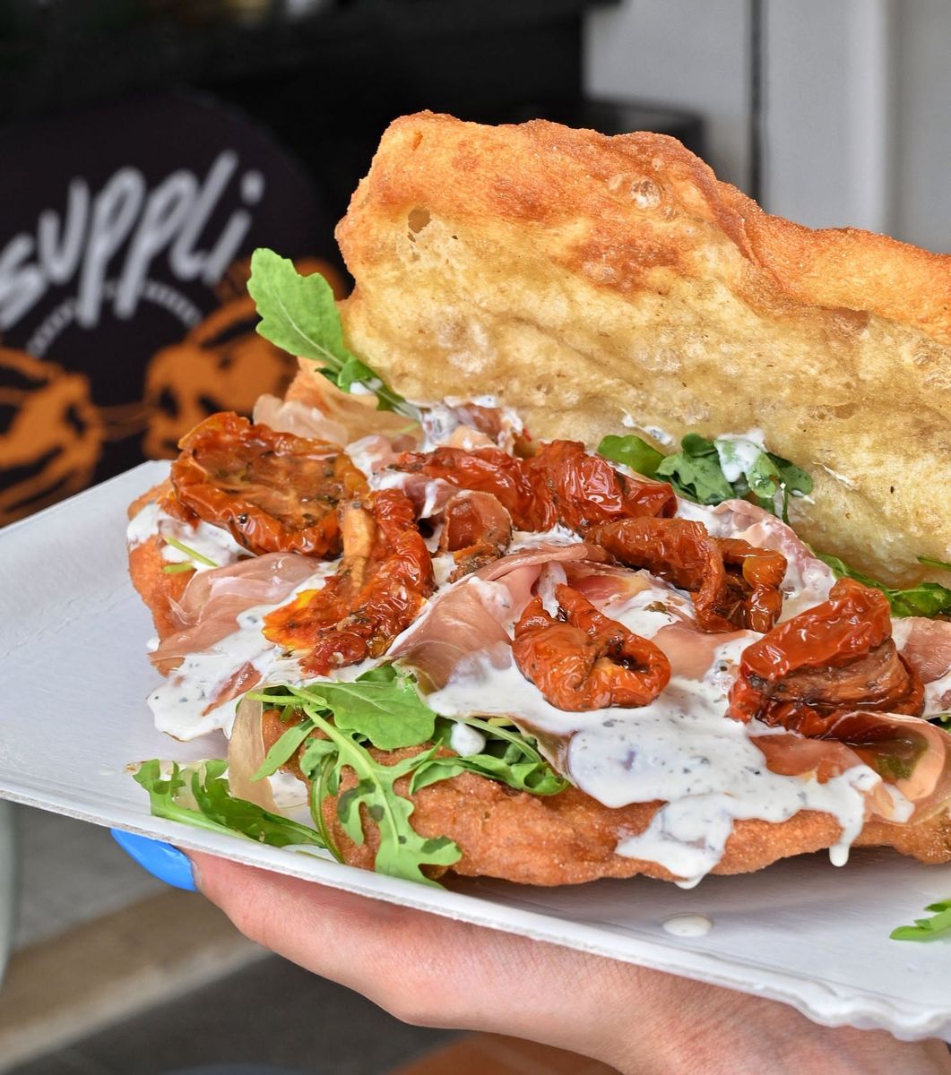 Una pizza fritta aperta tenuta in mano su un vassoio di carta: prosciutto crudo a fette, crema di burrata al tartufo e rucola, con l&rsquo;insegna arancione del locale sullo sfondo.