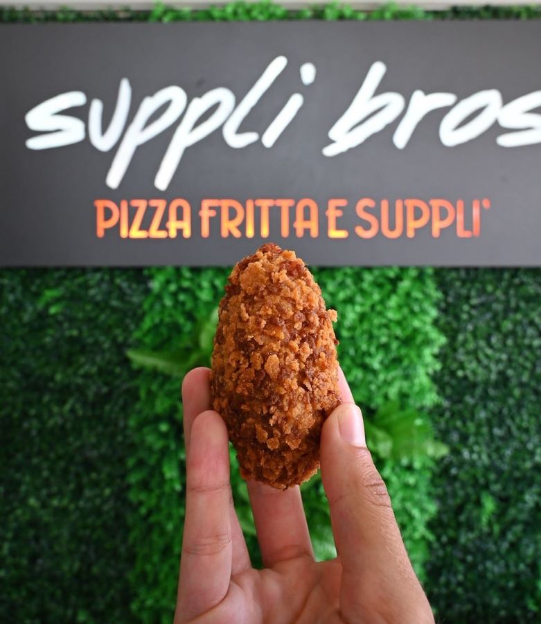 Un supplì appena fritto tenuto in mano davanti all&rsquo;insegna del locale, che porta la scritta Supplì Bros, pizza fritta e supplì.